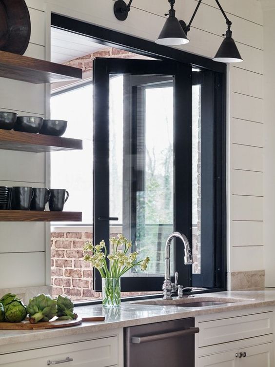 a white farmhouse kitchen with a black folding window, black sconces and thick open shelves with black tableware