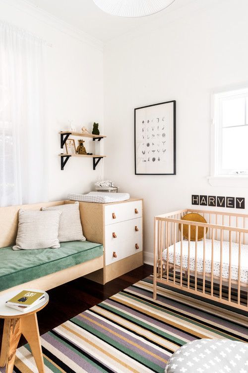 a Nordic chic nursery with white and stained furniture, a built in sofa, a brigth striped rug, open shelves and a printed pouf