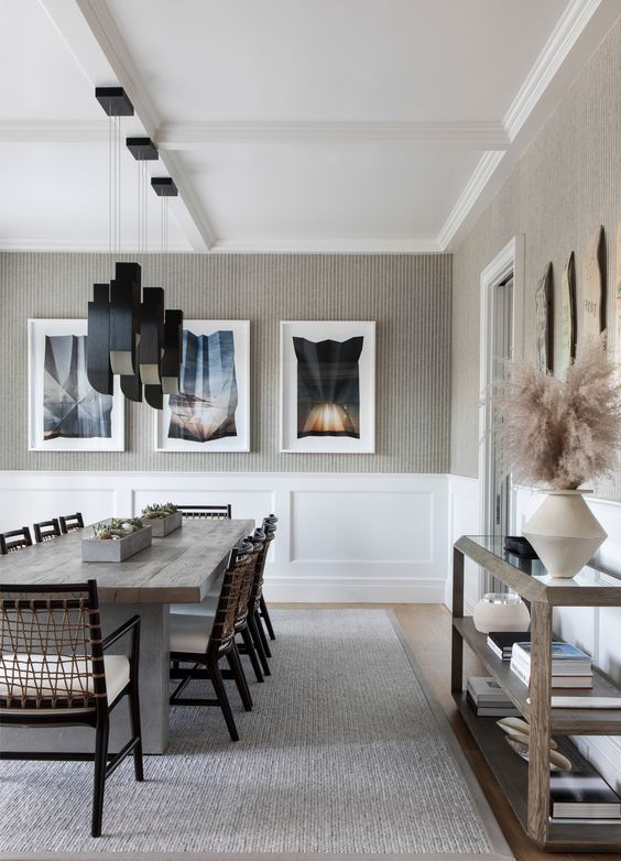 a bold and whimsy greige dining room with white paneling, a wooden table, woven chairs, black pendant lamps, a glass console table and gallery walls