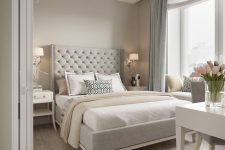 a chic greige bedroom with a grey upholstered bed, neutral bedding, a white vanity and a grey chair, blue curtains and some elegant lamps