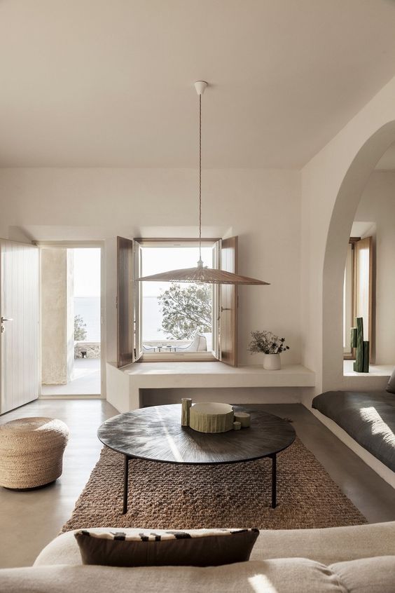 a chic greige living room with built-in sofas, a round metal coffee table, a pendant lamp, a jute pouf and a jute rug is very light-filled and welcoming