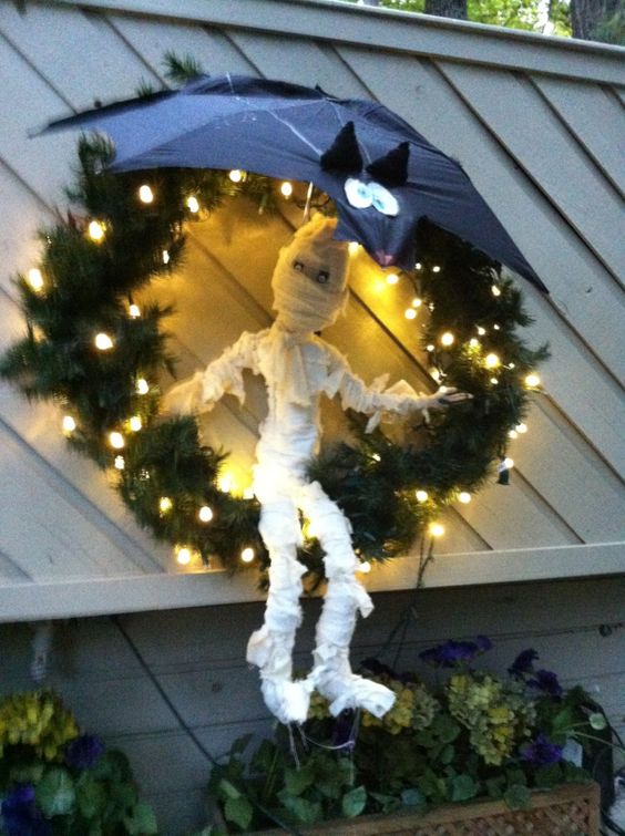 a funny Halloween mummy decoration on a Christmas wreath with lights and with a black bat on top to save it from rain