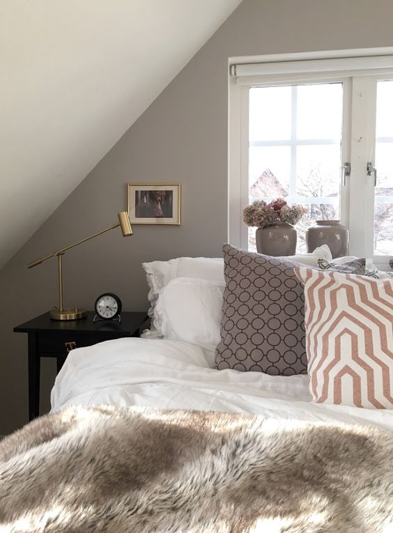 a greige attic bedroom with a large bed, printed pillows and faux fur, black nightstands and gold table lamp, greige vases and mini artworks