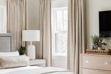 a greige bedroom with a grey upholstered bed, a stained dresser, beige curtains and an upholstered storage bench plus bedding