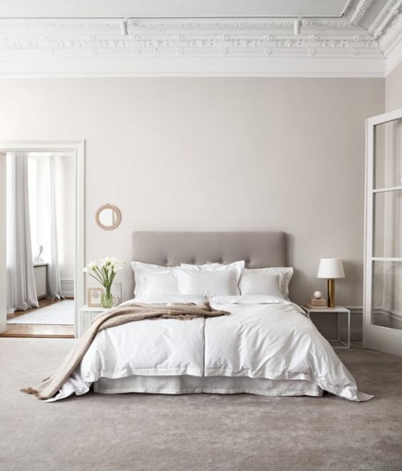 a greige bedroom with a grey upholstered bed, neutral bedding, a floor to ceiling mirror, matching white nightstands and table lamps