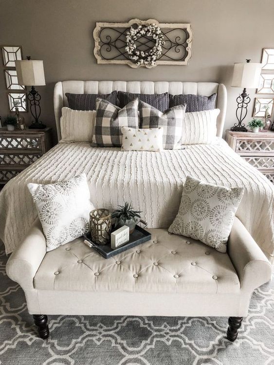 a greige farmhouse bedroom with neutral upholstered furniture, printed bedding, mirrored nightstands and table lamps, a cotton wreath