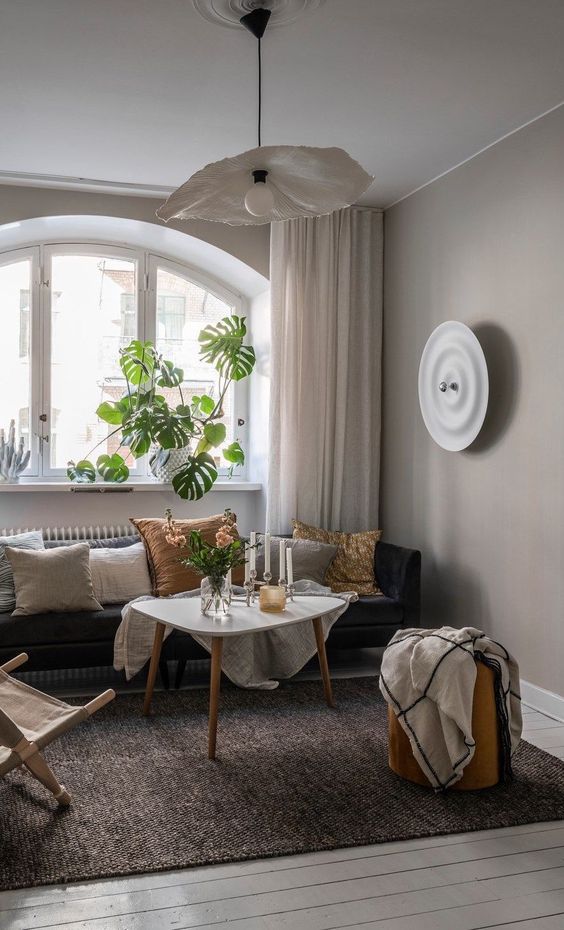 a greige living room with a black sofa, neutral and colorful pillows, a triangle table, a folding chair and a pendant lamp shaped as a flower