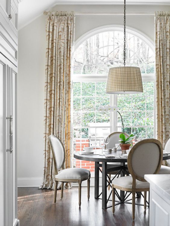 a lovely dining room with an oversized French window, a round table and rounded chairs, a pendant lamp and printed curtains