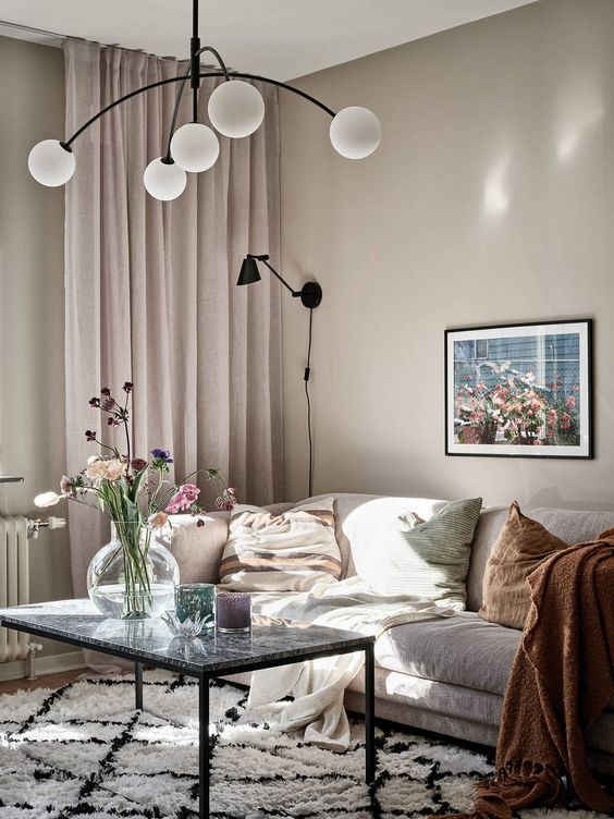 a lovely greige living room with a neutral sofa, a table with a stone tabletop, blush curtains, muted color pillows and a pretty chandelier