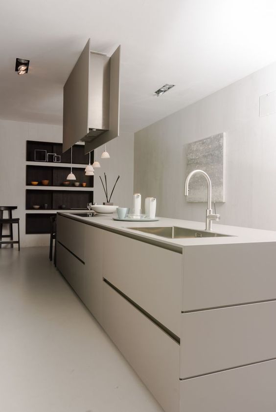 a minimalist greige kitchen with a sleek oversized kitchen island, a hood and hanging lamps plus built-in niches for tableware