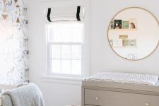 a neutral and pretty nursery with a wallpaper accent wall, greige furniture, a round mirror, ledges with books and an elephant basket for storage