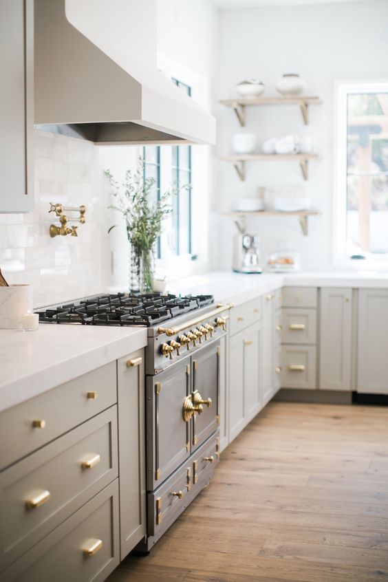 a pretty and refined greige kitchen with shaker cabinets, gold handles and white stone countertops, a white tile backsplash and open shelving