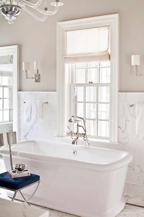 a refined greige bathroom with white marble tiles, a square tub, vintage fixtures and a beautiful chandelier plus a navy chair