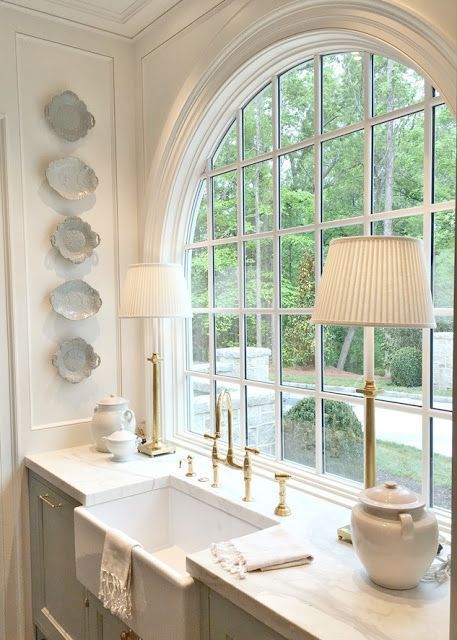 a refined neutral kitchen with grey cabinetry, white stone countertops, an arched French window, chic fixtures and lamps and decorative bowls on the wall