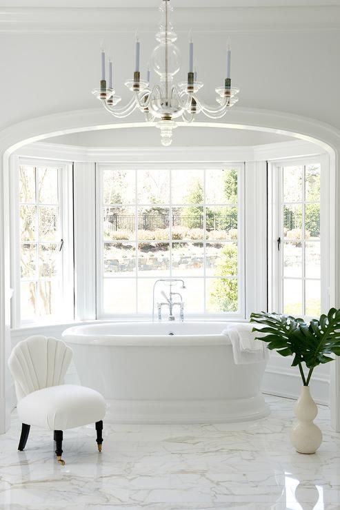 a refined white bathroom with a marble floor, an alcove with large French windows, a refined bathtub, a chair and a chic chandelier