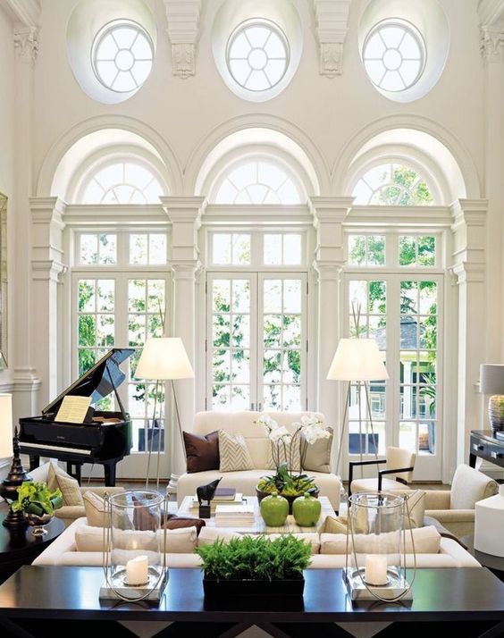 a sophisticated neutral living room with round and French windows, neutral furniture, floor lamps, a piano and a black console table