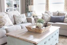 a vintage farmhouse greige living room with neutral sofas, a blue chest coffee table, an open storage bookcase, printed textiles