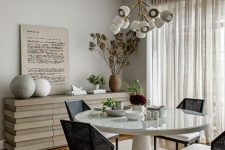an elegant and cool greige dining room with a catchy credenza, a round table, black chairs, a cool chandelier, some rocks and potted plants