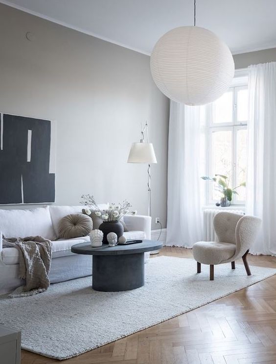 an ethereal greige living room with a white sofa and a cool wingback chair, a grey concrete round table, a floor and a pendant lamp