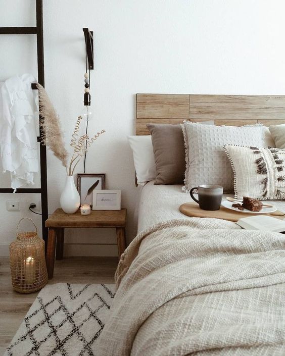 a neutral boho bedroom with a stained wooden bed, layered bedding, a wooden nightstand, candles, pampas grass and black touches