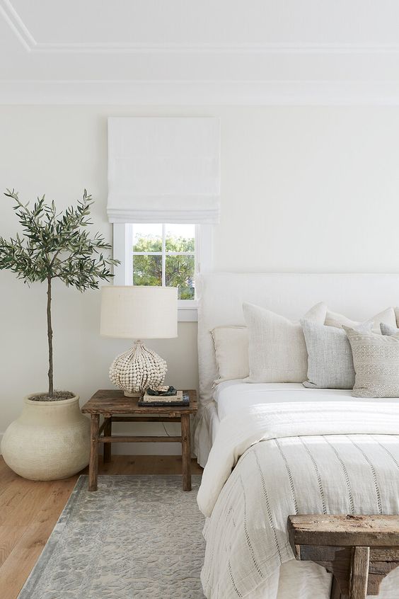 a beautiful all-neutral bedroom with a white upholstered bed, a wooden nightstand and a shabby chic bench, a potted plant and all neutral textiles