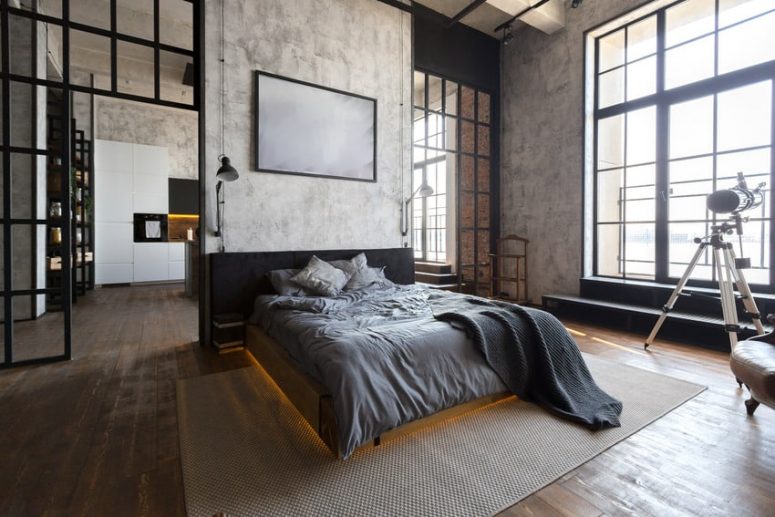 a chic industrial bedroom with concrete walls and a dark stained floor, a black floating bed with lights and grey bedding