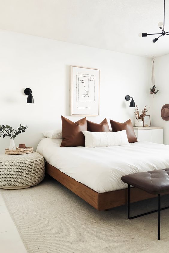 a chic mid-century modern bedroom in neutrals, with a rich-stained bed with neutral bedding, a leather bench, a woven ottoman and black sconces