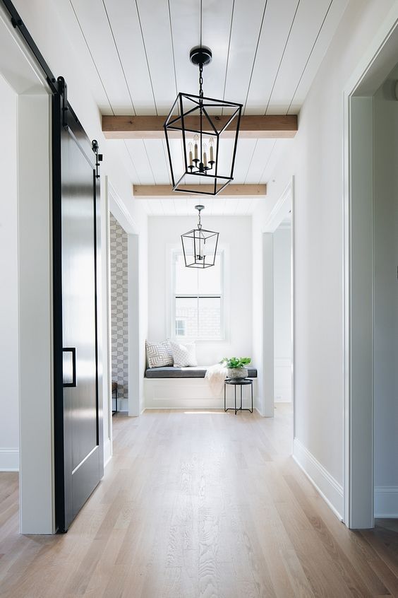 a light filled entryway with a white planked ceiling, stained wooden beams, a light stained floor, pendant lamps and a black sliding door