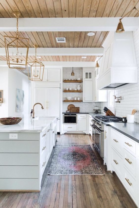 a white farmhouse kitchen with a light stained planked ceiling, a reclaimed wood floor, white cabinets and elegant gold pendant lamps