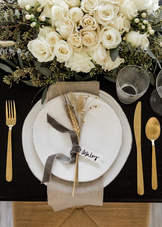 a gorgeous glam Thanksgiving table setting with a black uncovered table, white plates, gold cutlery, white florals and greenery and wheat
