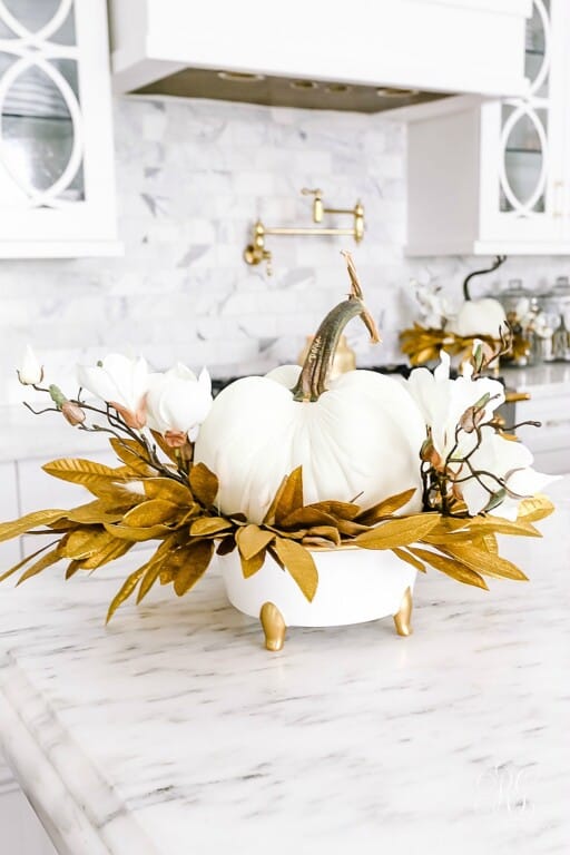 a glam Thanksgiving decoration of a white pumpkins, white blooms, gilded leaves on a white and gold stand is wow