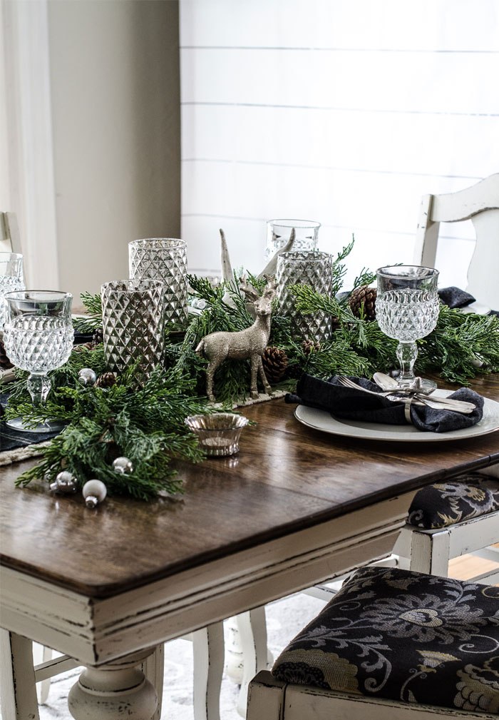 a forest glam Christmas tablescape with a greenery runner, silver ornaments, pinecones, glam and shiny candleholders, elegant glasses, dark napkins and silver chargers