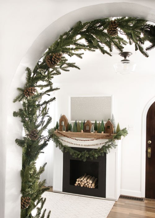 beautiful Scandi woodland Christmas decor with an evergreen and pinecone garland, wooden houses, garlands and mini trees on the mantel