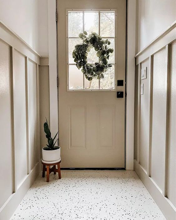 a beautiful greige entryway with wainscoting and a matching door, a white terrazzo floor and some greenery that enlivens the space
