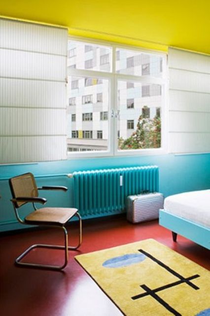 a bold bedroom with a yellow ceiling, turquoise walls and a bed plus a matching radiatoe, a red floor and a yellow rug