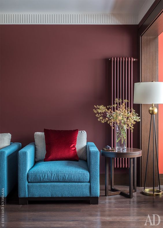 a bold living room with burgundy walls and a matching narrow radiator, bright blue chairs, a chic round side table and a floor lamp