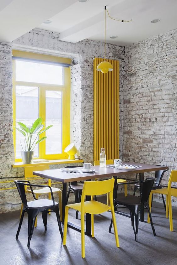 a catchy industrial dining room with brick walls, a dining table, black and yellow chairs, yellow window frames and a radiator on the wall