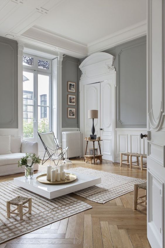 a jaw-dropping neutral Parisian space with grey walls, white wainscoting, crown molding, white furniture and neutral-colored chairs and stools