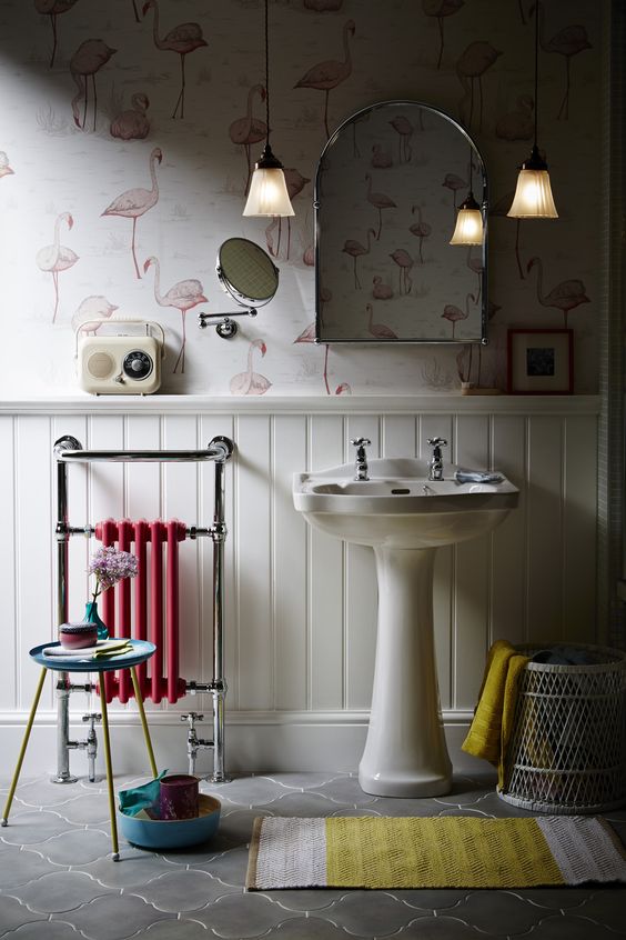 a lovely bathroom with flamingo printed wallpaper and white wainscoting, a free-standing sink, a small pink radiator, a bold rug and pendant lamps