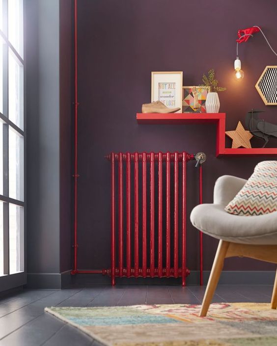 a modern living room with deep purple walls and a floor, a hot red radiator and a matching shelf for decor, a neutral chair and a printed pillow