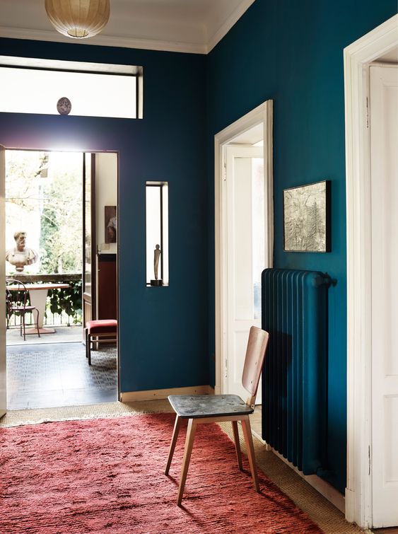 a navy entryway with white doors and framing, a matching navy radiator, a red rug and a simple chair is a chic space