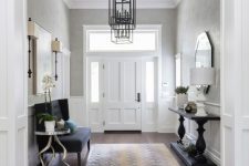 a sophisticated entryway with greige walls, white wainscoting and the door, a black upholstered bench, a black console table and some artworks and a mirror