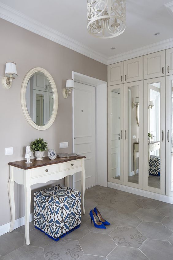 a vintage greige entryway with a creamy built-in storage unit with mirrors, a chic console table, an oval mirror, lamps and a printed pouf