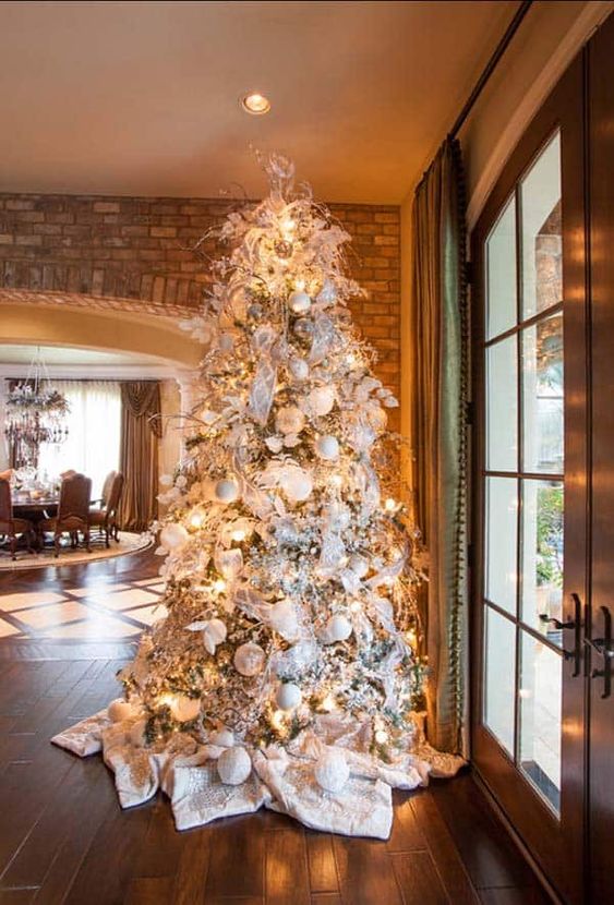 a winter wonderland Christmas tree with white and silver ornaments, lights, ribbons, branches and white blankets covering the base of the tree