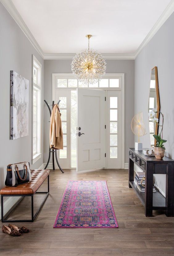 an airy and flowy entryway with greige walls, a leather upholstered bench, a black console, some art and a mirror with a catchy shape plus a colorful rug