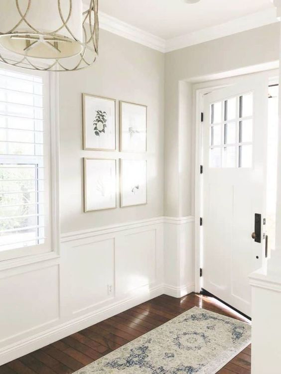 an airy and light-filled greige entryway with white wainscoting, a chandelier, a mini gallery wall and a white door