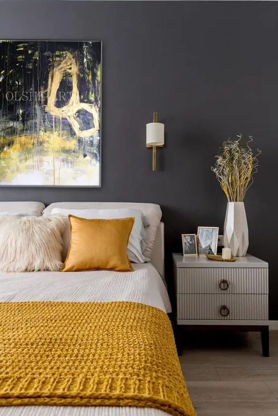 a bold bedroom with a graphite grey wall, creamy furniture, creamy and yellow bedding, a bold artwork over the bed