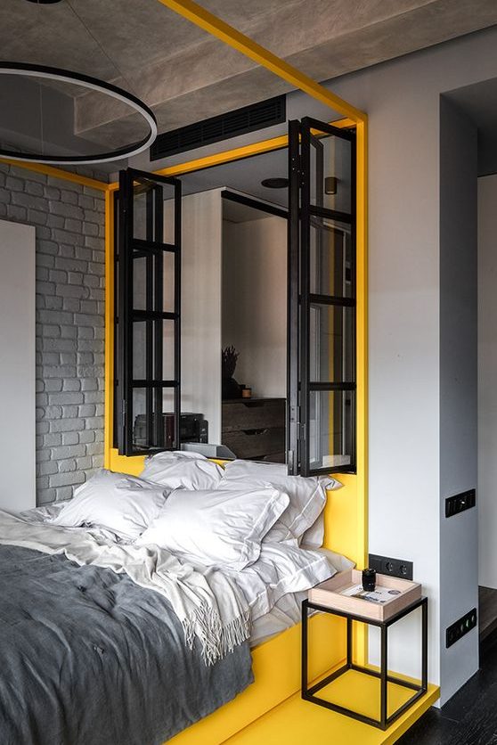 a modern industrial bedroom with light grey walls, a grey brick wall, a yellow zone with a window and a yellow bed with grey bedding