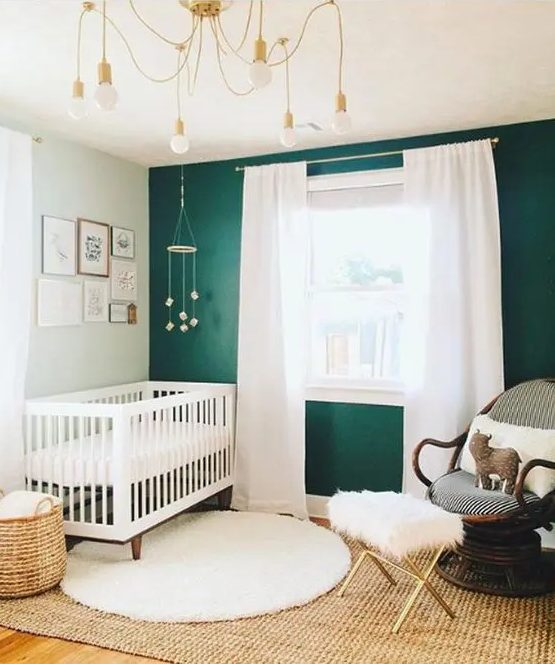 a boho modern nursery with a green accent wall, a white crib and a dark stained chair, neutral textiles, a whimsy chandelier and a basket for storage