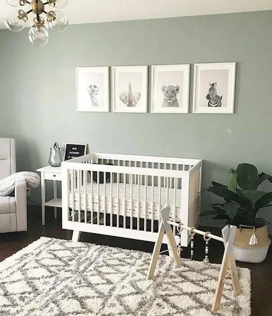 a cozy and cute nursery with a sage green accent wall, white furniture, a potted plant, a chic chandelier and a lovely printed rug